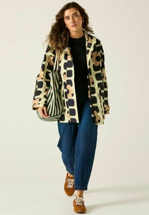Mujer caminando con una chaqueta beige y negra con estampado, vaqueros azules, zapatillas marrones y llevando un bolso de rayas verde y blanco.