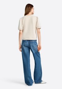 Haut beige à manches courtes avec des épaules bouffantes et une coupe décontractée, associé à un jean bleu à jambes larges avec poches arrière et délavage clair.