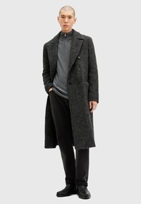 Grau wollener Mantel im Doppelreißverschluss-Design, präsentiert über einem grauen Kapuzenpullover und schwarzen Hosen, kombiniert mit schwarzen Stiefeletten.