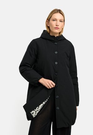 Kobieta ubrana w czarny płaszcz z kapturem i guzikami, częściowo otwarty, odsłaniający białą i czarną kropkowaną podszewkę.