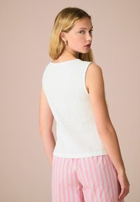 Top sans manches texturé blanc, encolure ronde. Le mannequin porte un pantalon rayé rose et blanc, vu de dos, avec des cheveux blonds doux.