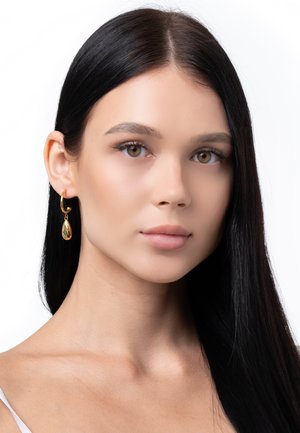 Goldene Tropfenohrringe mit einer glatten, polierten Oberfläche hängen an einem dünnen Ohrreif. Das elegante Design ergänzt das lange, gerade, dunkle Haar des Models.