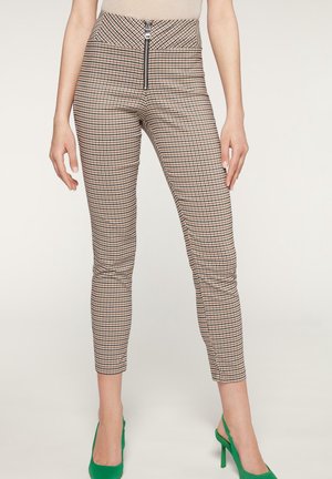 Nainen, jolla on päällään beige- ja ruskearuudulliset kapeat housut, joissa on vetoketju edessä, yhdistettynä kirkkaanvihreisiin korkeakorkoisiin slingback-kengin.