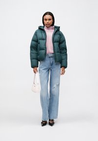 Casaco puffer verde com capuz, fecho éclair aberto, suéter rosa de malha, jeans claros rasgados azuis, e sapatos pretos de bico fino. Pequena mala branca.