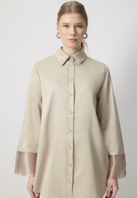 Chemise beige clair en tissu lisse, dotée d'une fermeture à boutons sur le devant, d'un col et de manches transparentes avec un subtil éclat.