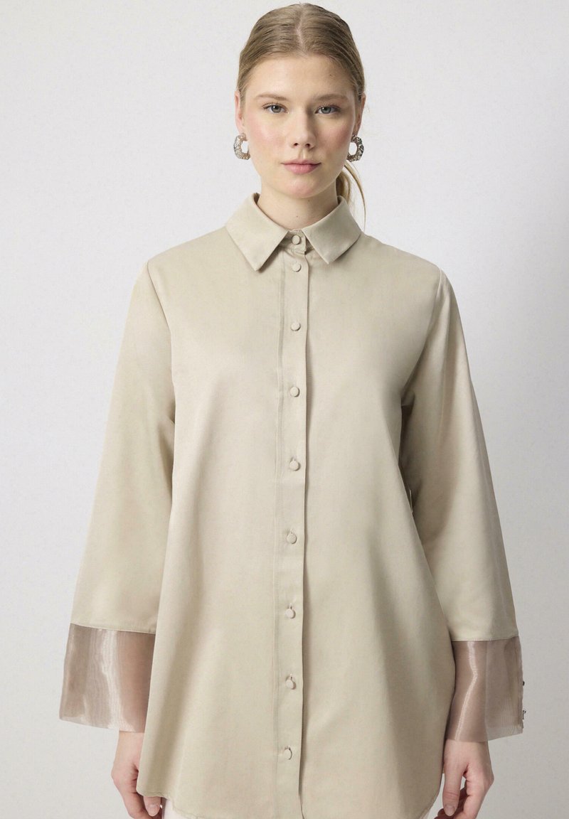 Chemise beige clair en tissu lisse, dotée d'une fermeture à boutons sur le devant, d'un col et de manches transparentes avec un subtil éclat.