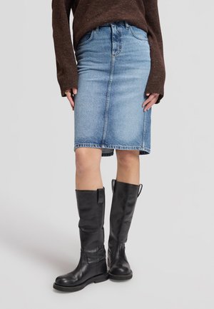 Personne portant une jupe en jean bleue mi-longue, des bottes noires montantes aux genoux et un pull marron à manches longues, debout sur un fond clair.
