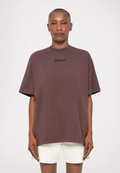 Camiseta de gran tamaño en color marrón chocolate con mangas cortas, cuello redondo y logo negro de "STIEGLITZ" en el pecho, combinada con pantalones cortos blancos.