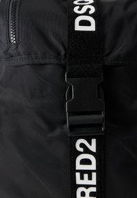 Μαύρη τσάντα από νάιλον με γυαλιστερό φινίρισμα, με φαρδιά λουράκι με λευκά γράμματα και πλαστική αγκράφα, καθώς και μικρή τσέπη με φερμουάρ.