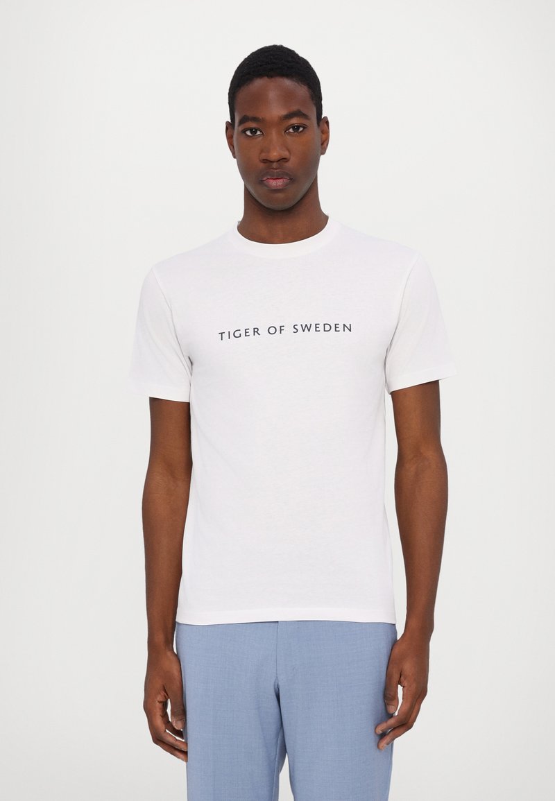 Weißes Baumwoll-T-Shirt mit dem Schriftzug "TIGER OF SWEDEN" in Marineblau. Kurze Ärmel und runder Halsausschnitt. Glatte Textur.