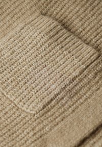 Beige strikket stof med en tekstureret overflade og en firkantet lomme. Vævningen viser en blanding af lyse og mørke fibre.