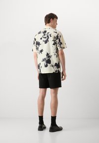 Camicia a maniche corte di colore crema con motivo floreale nero, abbinata a pantaloni neri. Indossata con calzature nere e calze, vista da dietro.