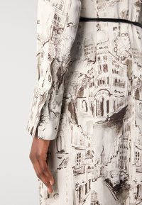 Long-sleeve dress featuring a cream fabric with brown architectural sketches, buttoned cuffs, and a fitted black waistband.