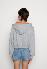Felpa grigia corta con un ampio cappuccio, foderata in arancione e con polsini elasticizzati, indossata su pantaloni denim blu con orli sfilacciati.