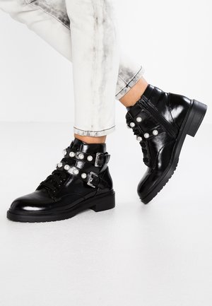 Czarne błyszczące botki za kostkę z paskami ozdobionymi perłami, z bocznymi zamkami, noszone z jasnoniebieskimi dżinsami podwiniętymi do góry na białym tle.