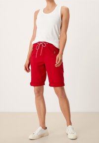 Pantalones cortos rojos con dobladillos enrollados, bolsillos laterales con cremalleras y cordón blanco. Combinados con una camiseta sin mangas blanca y zapatillas blancas.