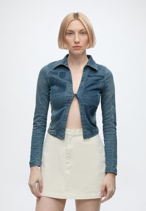 Jeune femme aux cheveux blonds courts porte une veste en jean bleue cintrée boutonnée une fois et une jupe blanche taille haute, debout sur un fond blanc.