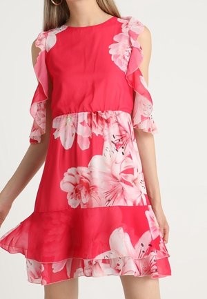 Rode bloemenjurk met open schouders, ruches aan mouwen en zoom, ingestikte taille, gedragen door een staand persoon met lang haar.