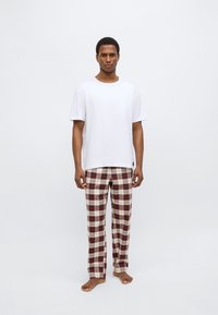 Weißes Baumwoll-T-Shirt mit kurzen Ärmeln, kombiniert mit karierten Schlafanzughosen in Burgunder- und Cremetönen. Das Model ist barfuß.