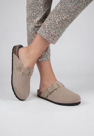 Füße, die beige Slip-On-Clogs mit Schnallen und schwarzen Sohlen tragen, kombiniert mit hellfarbenen gemusterten Hosen vor einfarbigem Hintergrund.