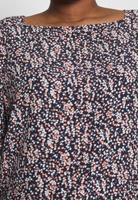 Blouse marine avec un décolleté carré, ornée d'un motif floral éparpillé en blanc, orange et bleu clair. Tissu doux au toucher texturé.