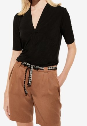 Kobieta w czarnej bluzce z krótkim rękawem i dekoltem w serek, włożonej do brązowych, wysokowysuniętych szortów z czarno-białym wzorzystym paskiem.