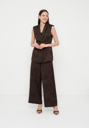 Γυναίκα χαμογελά, στέκεται με τα χέρια σταυρωμένα, φοράει αμάνικο σκούρο σακάκι με σχέδια, παντελόνια με φαρδιά πόδια και ψηλοτάκουνα ανοιχτά παπούτσια, σε λευκό φόντο.