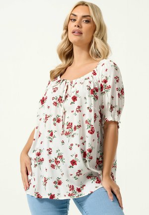 Blondynka ubrana w białą bluzkę z czerwonym kwiatowym wzorem i bufkami na rękawach, w połączeniu z jasnoniebieskimi dżinsami, pozująca na jednolitym tle.