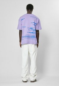 Lilac t-shirt with short sleeves, featuring a blue abstract pattern on the back. Paired with white pants, the outfit presents a casual look.