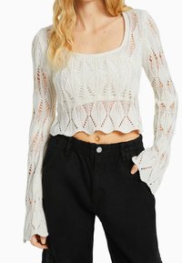 Witte gebreide crop top met lange, uitlopende mouwen voorzien van een bladvormig patroon en een gegolfde zoom, gecombineerd met zwarte cargo broek met trekkoord.