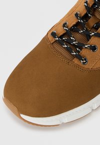 Zapatillas de ante marrón con punta redonda, cordones de patrón blanco y negro, y suela de goma blanca. Textura suave y detalles de costuras reforzadas.