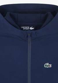 Tmavomodrá športová bunda s kapucňou, vybavená predným zipsom, sieťovanou podšívkou a malým zeleným logom krokodíla na hrudi.