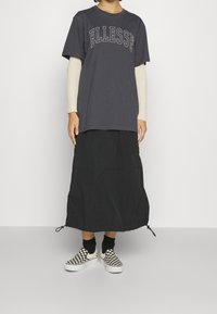 Camiseta gris con "ELLESSE" impresa en blanco, superpuesta a una blusa de manga larga con canalé en color crema. Falda negra con dobladillo ajustable y zapatos de cuadros tipo slip-on.