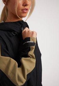 Svart och beige vattentät jacka med huva, med kontrasterande färgade ärmar och en grå dragkedja; textil med matte finish.
