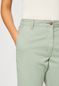 Osoba ubrana w jasne zielone spodnie z ręką w bocznej kieszeni oraz biały dziergany top włożony w pas.