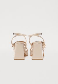 Des sandales à talons hauts en cuir verni nude présentent un talon bloc et un design de lanières croisées. La vue de dos met en avant des lanières de cheville réglables.