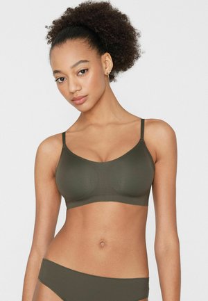 Mladá žena s kudrnatými vlasy, která má na sobě ladící olivově zelenou podprsenku a spodní prádlo, čelí přímo, na pozadí jednobarevné bílé zdi.