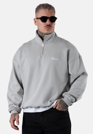 Pull-over à fermeture quart de zip gris clair en tissu texturé avec coupe ample, poignets côtelés et logo de marque brodé sur la poitrine. Lunettes de soleil noires portées.