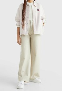 Light beige corduroy jacket with a patch logo, paired with high-waisted cream wide-leg pants and white sneakers. Smooth texture, relaxed fit.