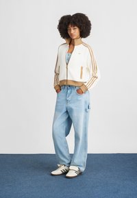 Casaco curto branco com riscas castanhas e fecho, combinado com jeans de perna larga azul-claro e sapatos brancos com pormenores pretos. Tecido texturizado.