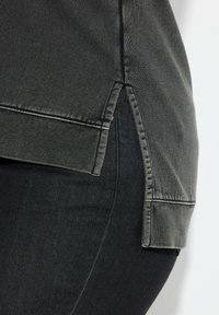 Camisa gris oscuro con una abertura lateral. Presenta una tela suave y texturizada con costuras contrastantes a lo largo del dobladillo. Combinada con vaqueros negros.