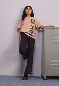 Beige Nike póló nagy nyomtatott logóval, fekete bővülő nadrággal párosítva. A modell egy szürke háttér előtt áll, egy szürke szekrény mellett.