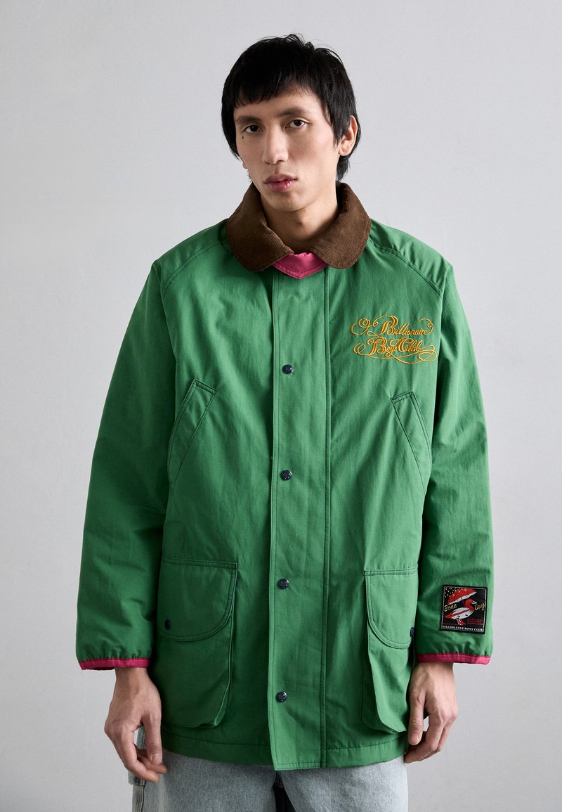 Grøn jakke med brunt kordfløjlskrage, der har to forlommer, knaplukning og logo-patch på ærmet. Struktureret stof.