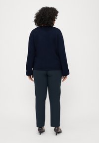 Pull à côtes bleu marine, coupe décontractée, manches longues, associé à un pantalon tailleur vert foncé, porté avec des chaussures à talons noires.