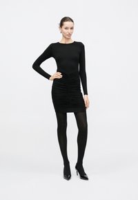 Γυναίκα με εφαρμοστό μαύρο μίνι φόρεμα με ραβδώσεις και μακριά μανίκια, μαύρο καλσόν και μαύρα ψηλοτάκουνα παπούτσια, στέκεται με το ένα χέρι στη μέση.