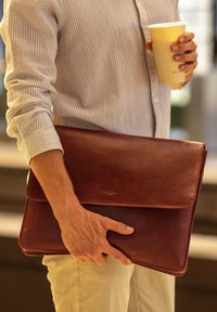Braune Lederlaptoptasche mit Klappenverschluss, glatter Textur und dezentem Branding. Der Halter trägt auch eine Kaffeetasse.
