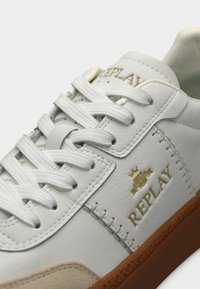 Λευκό δερμάτινο αθλητικό παπούτσι με χρυσό λογότυπο, υφασμάτινες μπεζ σουέντ λεπτομέρειες και λευκά κορδόνια. Διαθέτει καφέ λαστιχένια σόλα και ραμμένες λεπτομέρειες.