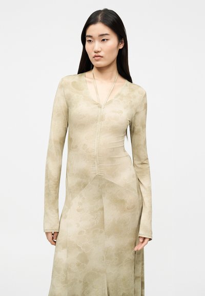 Νεαρή γυναίκα με μακριά σκούρα μαλλιά, φορώντας εφαρμοστό φόρεμα σε μπεζ χρώμα με μακριά μανίκια, ντεκολτέ σε σχήμα V και διακριτικές λεπτομέρειες ραφής.