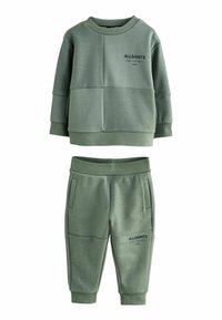 Olīvu zaļš bērnu treniņtērpa komplekts, kas sastāv no krekla ar apaļu kakla izgriezumu, uz kura ir teksts un strukturēti paneļi, kopā ar biksēm ar elastīgu jostu.