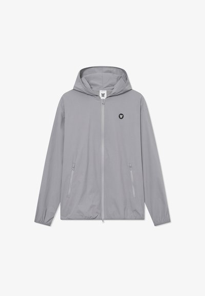 Veste grise clair avec capuche, fermeture éclair à l'avant, deux poches latérales et un petit logo noir sur la poitrine. Fabriquée en tissu léger et lisse.
